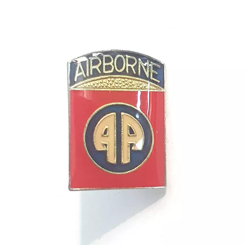 Pin's bouclier titré "AIRBORNE" sur un bandeau noir et illustré avec un logo "AA" (All American) en lettres stylisées.